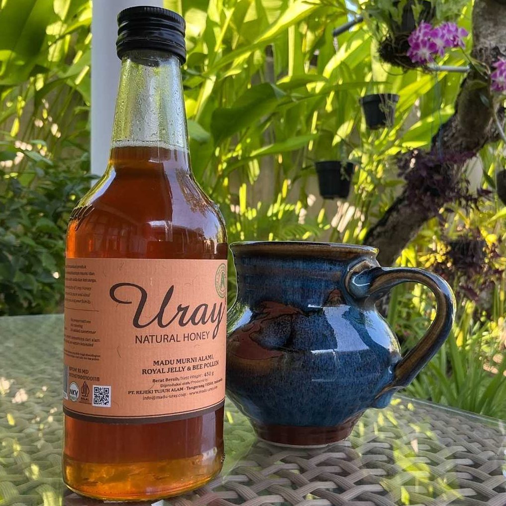 A bottle of amber colored honey next to a blue ceramic mug set a against a lush tropical garden