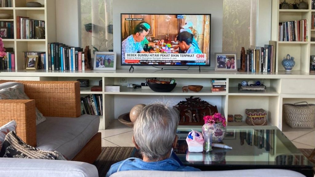 My mother with grey hair watching tv in her living room in Indonesia